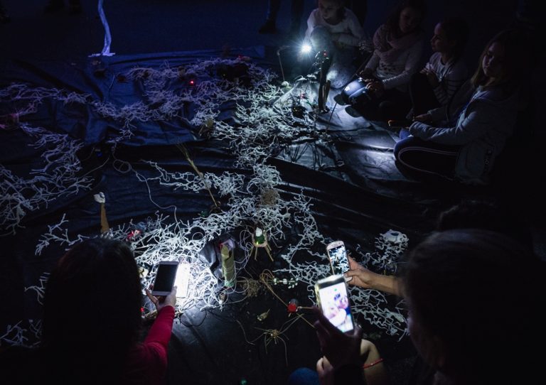 Grupa ludzi siedzi w ciemnym pomieszczeniu wokół instalacji artystycznej wykonanej z poplątanych białych przewodów lub sznurków na czarnym materiale, używając swoich telefonów jako latarek i robiąc zdjęcia ekspozycji.