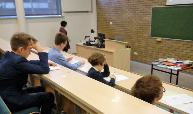 Kilku uczniów biorących udział w konkursie przyrodniczym siedzi w sali wykładowej, pisząc na kartkach, podczas gdy nauczyciel stoi z przodu przy biurku i tablicy. Książki leżą na pobliskim stole. Sala ma ceglaną ścianę i duże okna.