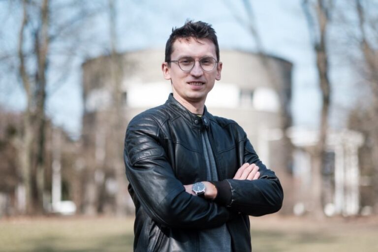 Młody mężczyzna uśmiecha się, w tle budynek, drzewa.